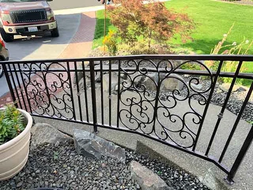 An intricately designed ornamental iron works railing along a garden path. The railing showcases detailed scrollwork and decorative patterns, demonstrating the fine craftsmanship of iron works ornamental. The dark wrought iron contrasts beautifully with the greenery and landscaping, enhancing the visual appeal of the garden. The path is lined with rocks and plants, leading up to a well-maintained lawn and driveway. This setup highlights the use of railings wrought iron in outdoor spaces, providing both safety and aesthetic enhancement to garden paths and residential landscapes.