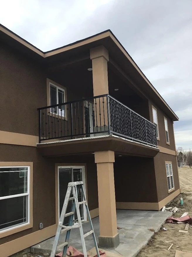 A two-story home featuring a balcony with deck railings metal. The railings are designed with vertical bars and decorative elements, emphasizing the artistic touch of ornamental iron works. The house structure includes prominent support beams, indicative of construction steel beam usage, ensuring stability and durability. The balcony provides a safe and stylish outdoor space, seamlessly integrating with the building's modern design. A ladder in the foreground suggests ongoing work, possibly related to on site welding or welding repair services. This setup highlights the practical application of fabricated structural steel and the craftsmanship involved in creating superior fence and railing installations.