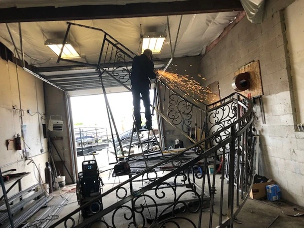 A skilled worker performing on site welding on a beautifully crafted ornamental iron works staircase railing in a workshop. Sparks fly as the welder uses specialized equipment to ensure the railing's structural integrity and aesthetic finish. The railing features intricate scrollwork, typical of iron works ornamental, which adds both elegance and durability. The workshop is equipped with various metalworking tools and materials, indicative of the environment where structural iron and steel workers operate. This setup demonstrates the blend of artistry and technical expertise required in welding repair services and fabricated structural steel projects, highlighting the meticulous process of creating high-quality iron railings for residential or commercial properties.