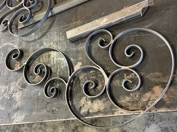 Detailed scrollwork pieces in the process of fabricated structural steel within a workshop. These decorative elements showcase the intricate craftsmanship typical of ornamental iron works and iron works ornamental. The scrolls are meticulously shaped and ready to be incorporated into larger designs such as gates, railings, or other decorative structures. The workshop environment, complete with metalworking tools and a workspace covered in metal shavings, highlights the precision and skill involved in creating high-quality railings wrought iron and other ornamental metal pieces. This setup underscores the artistry and technical expertise required in structural steelwork fabrication and metal pipe welding to produce aesthetically pleasing and durable iron works.