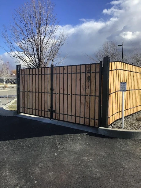 A solid fence constructed with vertical wooden panels secured within a sturdy black metal frame. The fence includes a wide double gate, indicating an entry point to a restricted or private area. The surrounding area features a paved asphalt surface and gravel landscaping, with a leafless tree and a cloudy sky in the background. This fence combines the natural aesthetic of wood with the durability and strength of metal, showcasing a high-quality superior fence and railing installation suitable for both residential and commercial properties.